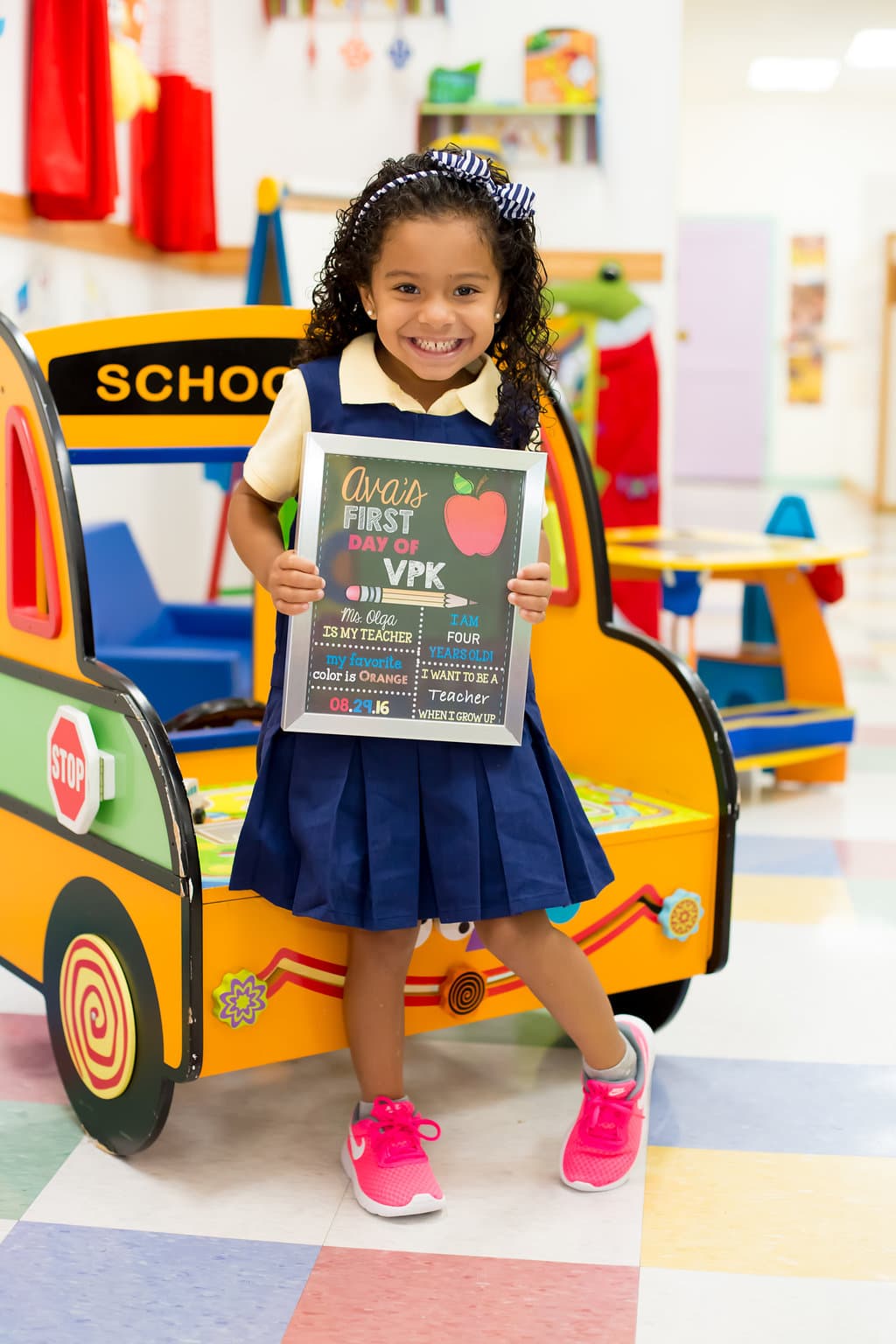 A smiling student holding a first-day-of-VPK sign beside the classroom school-bus play structure.